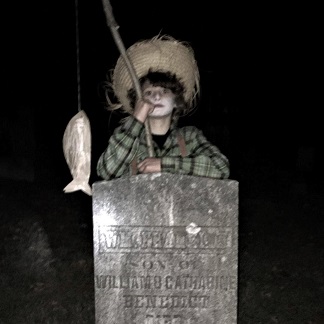 boy dressed as a ghostly boy from the past with a fishing pole, leaning on a headstone in cemetary