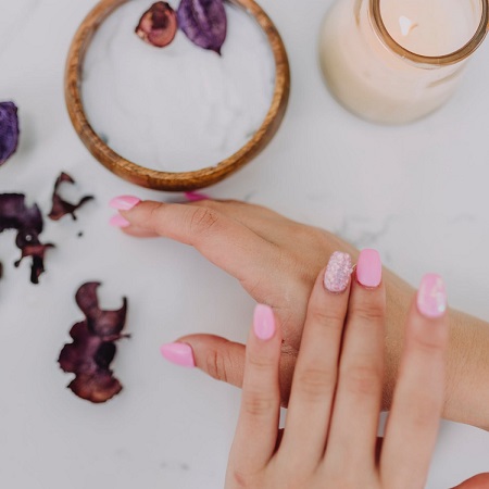 hands with pink nails rubbing in body scrub from small container nearby