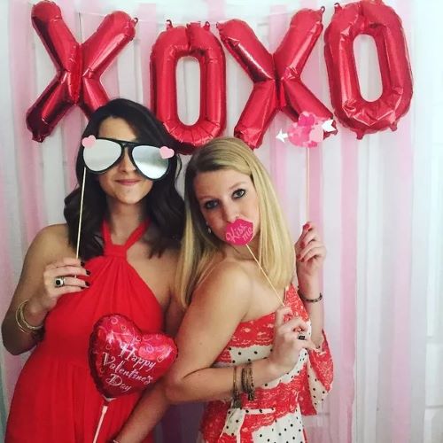 two teen girls dressed in red with xoxo banner hanging above and funny sunglass and lip cutouts on sticks