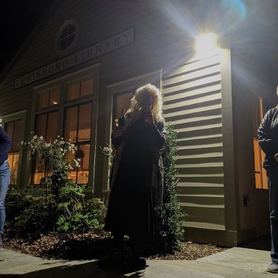 Woman in costume outdoors at night beneath Lewisboro Library sign.