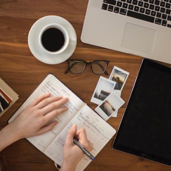 person writing in notebook alongside of laptop, cup of coffee, glasses