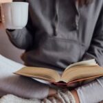 Someone sitting cross legged reading a book on lap while holding a mug