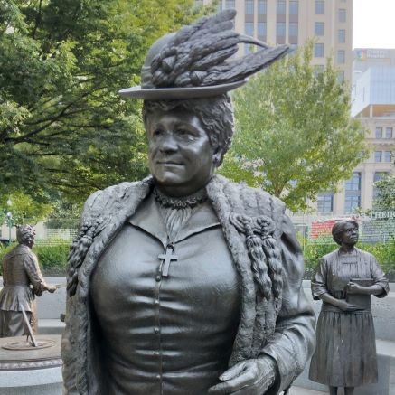 outdoor bronze stature depicting woman in a feathered hat, fur coat and old fashioned dress, wearing a cross