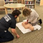 woman administering cpr on dummy as ems worker looks on