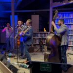 4 men on stage in front of microphones, one playing violin, another playing cello, another playing guitar and last man playing bass