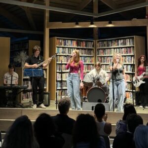 teen band on library stage: male keyboardist, male bass player, two female singers, male drummer and female guitarist