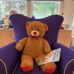 Stuffed Teddy Bear sitting in purple easy chair with paint brush under one paw and a book under the other paw.