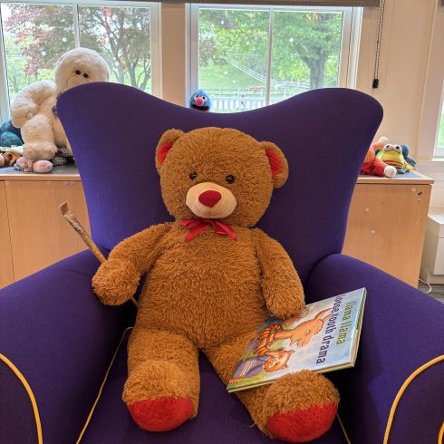 Stuffed Teddy Bear sitting in purple easy chair with paint brush under one paw and a book under the other paw.