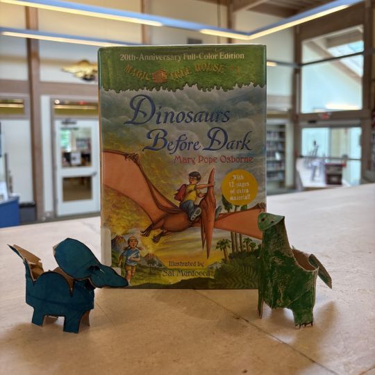 The book "Dinosaurs Before Dark" on shelf next to two dinosaurs created from cardboard.