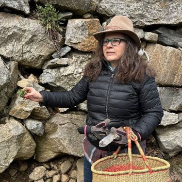 Woman outdoors by stone wall, displaying a plant she just dug up.