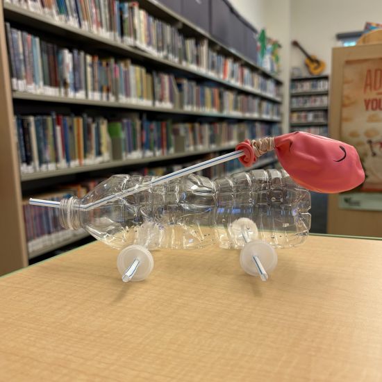 "car" made from empty water bottle, with plastic cap wheels and drinking straw axle. Drinking straw attached to balloon punctures the bottle to be blown into, to "power" the car