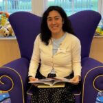 young woman in a yellow cardigan sitting on oversized purple easy chair, reading a children's book