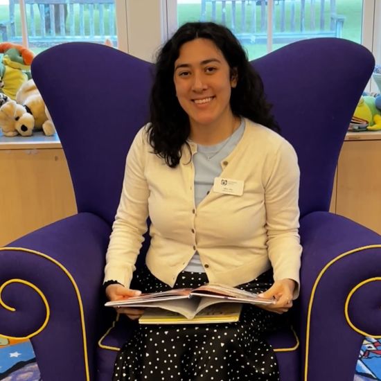young woman in a yellow cardigan sitting on oversized purple easy chair, reading a children's book