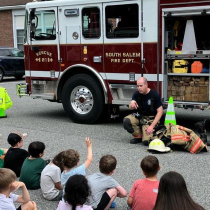 fireman seated in front of firetruck taking questions from children sitting on ground before him