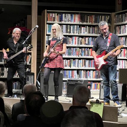 stage containing grey haired man playing bass guitar, woman with long blonde hair on guitar and second grey haired man playing guitar