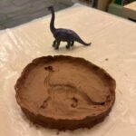 Toy dinosaur beside circle of brown clay with impression of dinosaur in it.