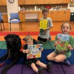 three children holding up picture books while sitting alongside a black dog