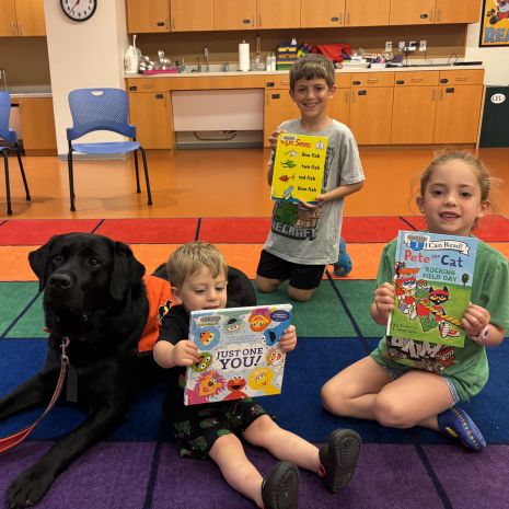 three children holding up picture books while sitting alongside a black dog