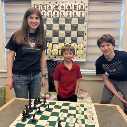 two teens alongside a young boy, in front of a giant chess board