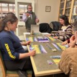 adults sitting at a table around a board game, as a male instructor explains the rules