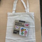 plain white tote bag laying flat on table with package of fabric markers on top