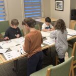 Children gathered around a table lined with paper, drawing routes for ozobot robots