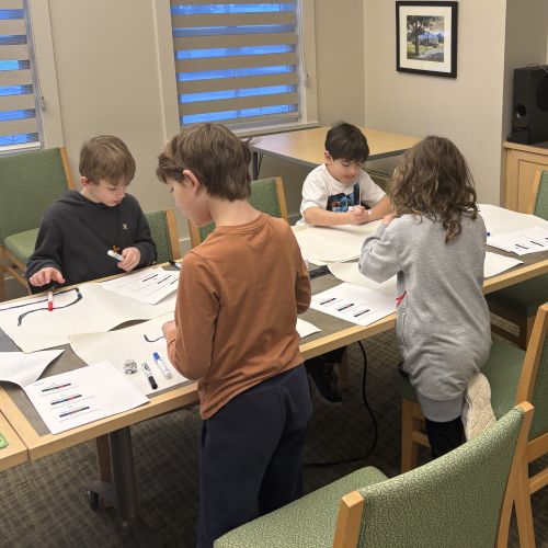Children gathered around a table lined with paper, drawing routes for ozobot robots