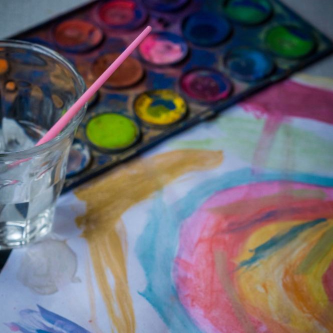 children's paint palette beside glass of water with small paintbrush in it, and painting of a rainbow