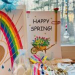 Hand drawn greeting card featuring a wheelbarrow filled with flowers, next to a beaded suncatcher hanging in window.