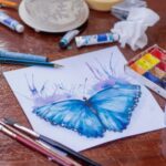 An image of a painting of a blue butterfly on a table surrounded by paints and brushes.