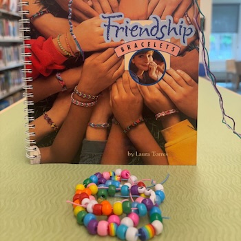 A stack of bracelets made out of colorful beads in front of a book about Friendship Bracelet making.