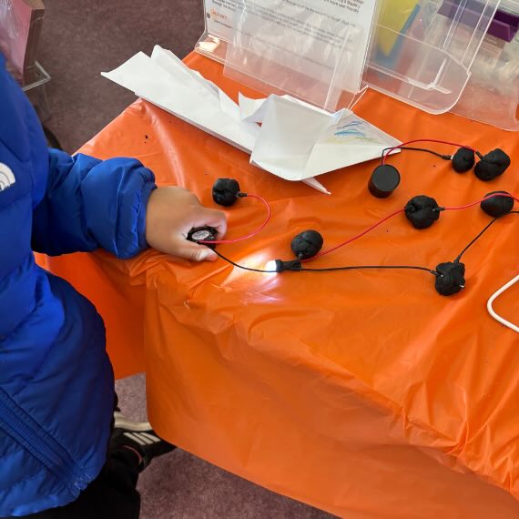 A small hand plays with a circuit project sitting on a table that has a lit up lightbulb.