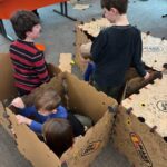 Four young children playing in a structure built by cardboard panels.