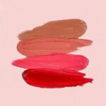 Four shades of red and brown lip gloss smeared on a pink background.