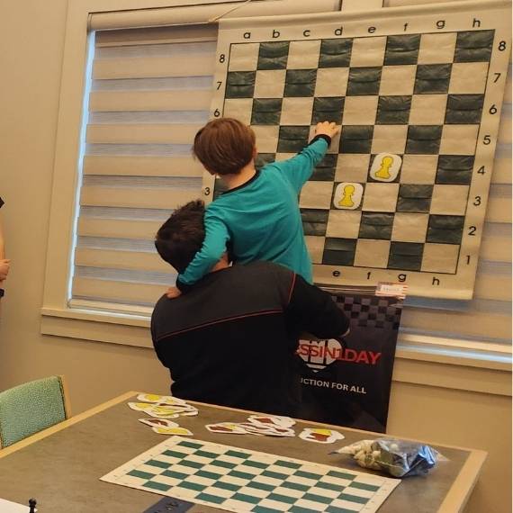 Child held up by adult as child touches a large cloth chess board hanging on the wall.