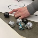 Small robots on a table with a hand drawing a line on a piece of paper.