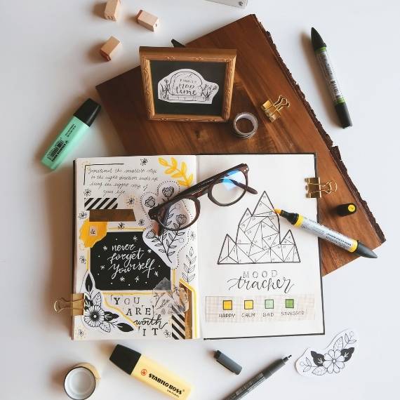A journal with pens and a pair of glasses on top a wooden panel sitting on a table