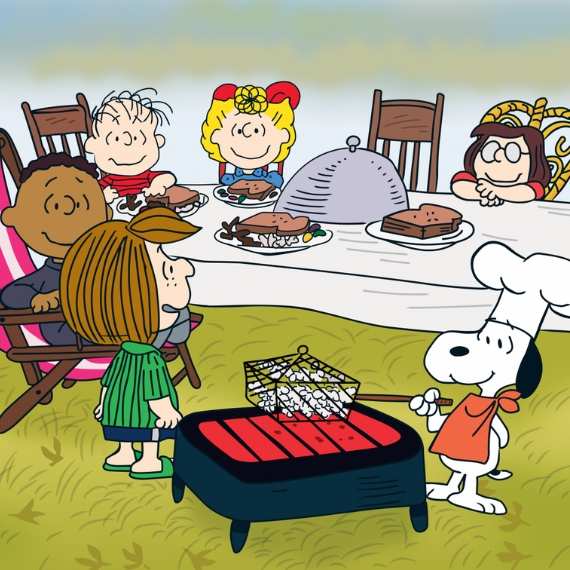 Cartoon image from PEANUTS of various children sitting around a table with food as a white dog in a chefs hat cooks popcorn.